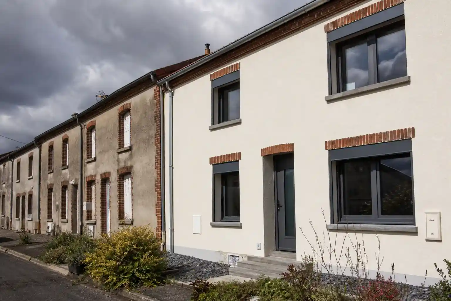 Alignement de maisons ouvrières du bassin minier à Cagnac-les-Mines, l’une rénovée avec enduit clair et menuiseries neuves, ciel changeant