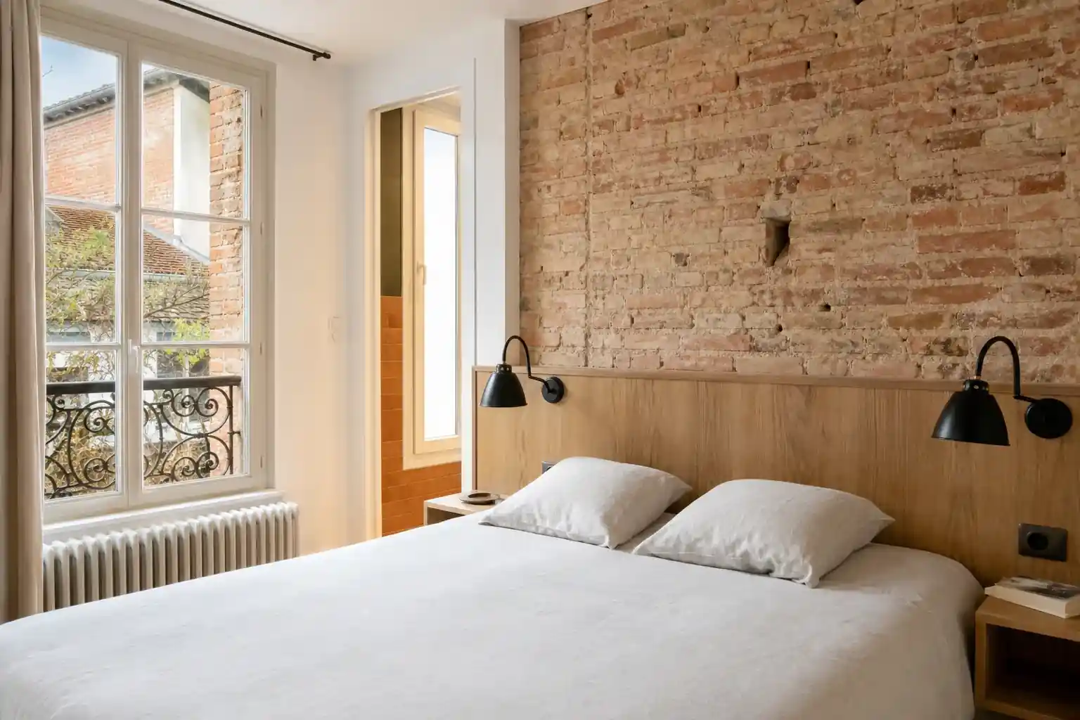 Chambre d’appartement rénovée avec mur de brique apparente conservé, tête de lit menuisée, luminaires appliques