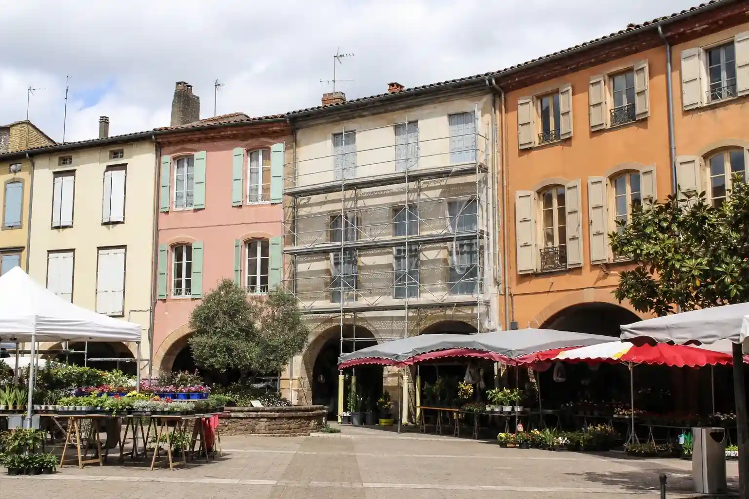 Place à arcades de la bastide de Réalmont, maisons de bourg colorées, une façade en cours de ravalement avec échafaudage discret, marché en fin de matinée