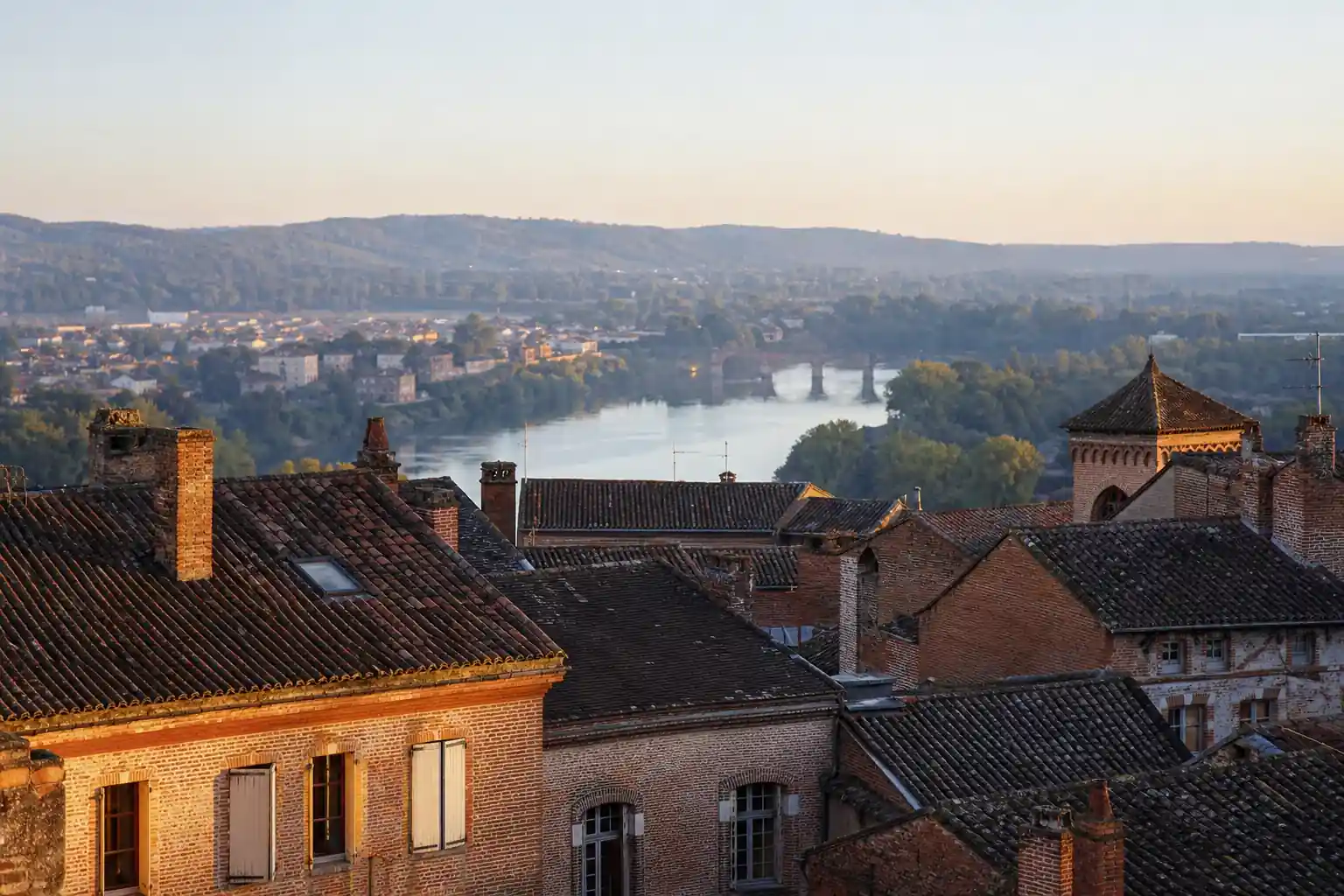 Vue de la vallée du Tarn autour d’Albi au petit matin, toits de tuile et façades de brique au premier plan, coteaux et villages alentour au loin